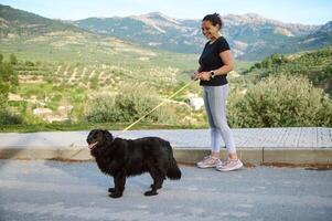 Side portrait of a woman in sportswear, walking her dog on leash in the nature. Multi ethnic enjoying walk with her pedigree purebred black cocker spaniel dog in the mountains nature outdoors photo