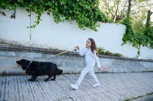 Happy child girl enjoying walking her dog outdoors in the nature. Pretty school age kid taking her pedigree black cocker spaniel dog for a walk on sunny day. Side view photo