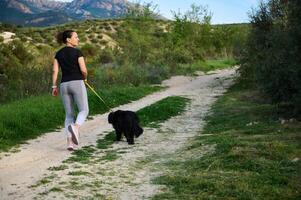 Full size shot of a happy woman smiling looking away, walking her dog in mountains nature outdoors. Rear view photo