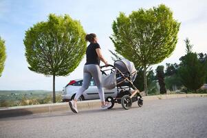 posterior ver de un joven mujer, joven madre corriendo trotar hacer ejercicio al aire libre con el paseante de su bebé a permanecer en forma después parto. foto