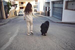 posterior ver de un bonito niña caminando su perro en Correa en el calle en soleado día. negro cocker spaniel mascota siendo caminado al aire libre. personas y animales concepto foto
