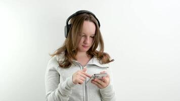Suche auf das Telefon zum geeignet Musik- im das Wiedergabeliste Teenager Mädchen im Kopfhörer wählt ein Melodie zu Hör mal zu Wind Sportbekleidung auf ein Weiß Hintergrund video