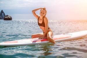 mujer cenar mar. Deportes niña en un tabla de surf en el mar en un soleado verano día. en un negro baños traje, él se sienta en un sapa en el mar. descanso en el mar. foto