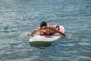 mujer savia mar. Deportes niña en un tabla de surf en el mar en un soleado verano día. en un negro baños traje, él mentiras en un savia en el mar. descanso en el mar. foto