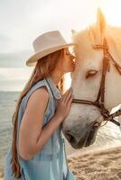 A woman in a dress stands next to a white horse on a beach, with photo