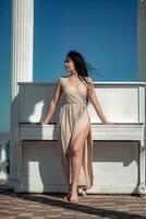 A woman in a long dress stands in front of a piano. The piano is white and has a few keys visible. The woman is posing for a photo, and the overall mood of the image is calm and serene. photo