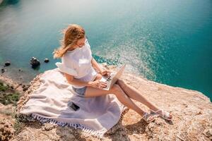 Freelance women sea working on the computer. Good looking middle aged woman typing on a laptop keyboard outdoors with a beautiful sea view. The concept of remote work. photo