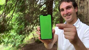 un hermoso Español hombre muestra un teléfono en cuales un verde pantalla chromakey él soportes en un bosque vistiendo un blanco camisa video