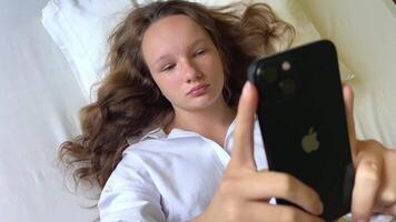 en flicka i en vit skjorta en tonåring lögner på en vit säng i henne händer hon innehar en svart iphone 13 hon utseende på de skärm lugnt höjning henne händer upp video