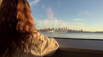 The fair-haired Girl sits with her back to the camera and looks out the window of sea bus. She sees Vancouver in front of her with tall houses We can also see her face reflected in the clear glass. video