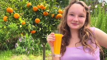 une adolescent fille des stands avec une verre de Orange jus sur le Contexte de une agrumes arbre mandarines ou des oranges pendre sur une arbre. elle sourit et regards dans le Cadre pouvez être utilisé à afficher jus video