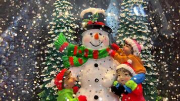 Snow globe with snowman stands between two snow-covered Christmas trees Near him are 3 children who just built him he has a black hat with a green scarf with red stripes Silvery snowflakes fly around video