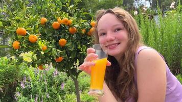 en Tonårs flicka på de bakgrund av ett orange träd i henne händer med en glas av orange juice blinkar in i de ram och gör ansikten video