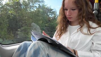 European girl with blond long hair in a white shirt rides in skytrain teenage girl diligently does her homework to hold a notebook on her knees through the window and goes home she is 13-15 years old video