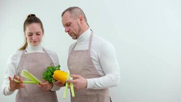 schort twee mensen familie liefde vegetarisme wit achtergrond Koken Cursus mannetje chef in koken uniform geeft les jong vrouw Koken klasse studenten hoe naar koken groenten mengen voedsel ingrediënten in keuken. video