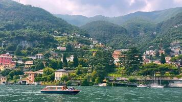 Scenic View of Lake Como with Lush Green Hills, A picturesque image of Lake Como, showcasing vibrant green hills, colorful lakeside houses, and leisure boats under a cloudy sky, capturing the serene video