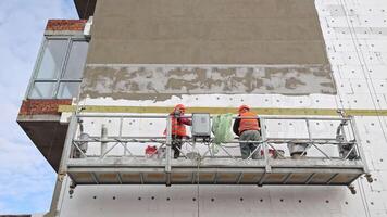 construcción trabajadores aislante edificio fachada, dos construcción trabajadores en reflexivo chalecos trabajando en andamio, aplicando aislamiento a un del edificio fachada. video