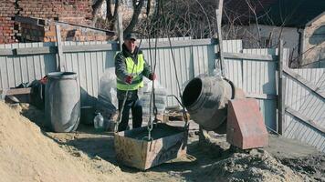 arbetstagare på konstruktion webbplats framställning cement, en konstruktion arbetstagare i en hög synlighet väst och keps manuellt blandning cement på en byggnad webbplats, omgiven förbi konstruktion material. video