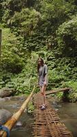 Girl sits on a bamboo suspension bridge over a small river, vertical video