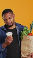 Vertical Young adult enjoying a cup of coffee during his grocery shopping spree, carrying a paper bag full of organic produce in studio. Man serving caffeine refreshment, zero waste. Camera B. video