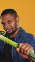 Vertical Pleased young adult pretending to start a fight with a cucumber and a leek, acting silly having fun in studio. Vegan guy playing around with natural eco friendly green vegetables. Camera A. video