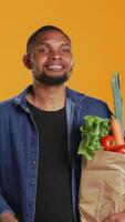 Vertical Cheerful person juggling with a green ripe apple on camera, carrying paper bag full of organic bio groceries. Smiling young man playing around with a fruit, sustainable lifestyle. Camera B. video