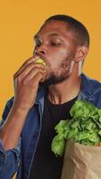 Vertical African american vegan guy taking a bite from a ripe natural green apple, eating freshly harvested organic fruits in studio. Person advertising local farming and ethically sourced goods. Camera A. video