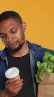 vertical joven adulto disfrutando un taza de café durante su tienda de comestibles compras juerga, que lleva un papel bolso lleno de orgánico Produce en estudio. hombre servicio cafeína refresco, cero desperdiciar. cámara una. video