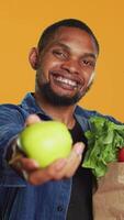 Vertikale afrikanisch amerikanisch Mann präsentieren ein frisch geerntet Grün Apfel, zeigen das organisch bio Früchte im Vorderseite von das Kamera. glücklich Person befürworten zum gesund Essen und vegan Ernährung. Kamera a. video