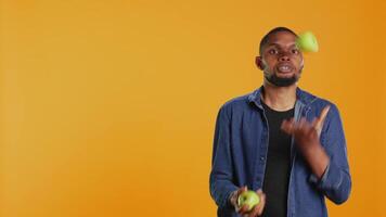 Playful person juggling in the studio with ripe green apples, keeping it in the air with continuous motion by throwing and catching. Confident skilled guy having fun with organic fruits. Camera B. video