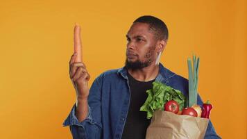 Vegan guy looking closely at a carrot to ensure its best shape, supporting local farming with ethically sourced produce. Young man carrying paper bag full of organic groceries. Camera A. video