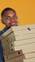 vertical confidente masculino mensajero que lleva grande pila de Pizza cajas a entregar rápido comida orden a clientela, para llevar Servicio en estudio. joven sonriente repartidor trayendo eliminar comida pila. cámara una. video