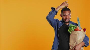 Young adult trying to keep balance with an apple on his head in studio, playing around and advocating for zero waste and sustainable lifestyle. Model carries paper bag with bio goods. Camera B. video