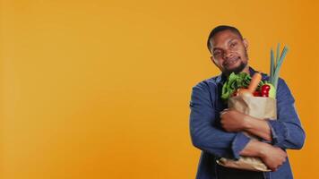 masculino modelo abraçando dele papel saco cheio do orgânico caseiro comida, desfrutando pantanoso comendo a partir de local bio supermercado. jovem vegano pessoa recomendando eticamente originado produzir. Câmera b. video