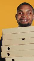 Vertical African american pizzeria deliveryman holding stack of pizza boxes to deliver food order to customer in studio. Friendly takeout service employee carries takeaway meal package. Camera A. video