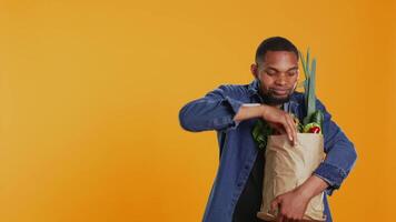 veganistisch volwassen presenteren een rijp wortel van zijn boodschappen papier tas, aanbevelen lokaal landbouw met etisch afkomstig eco vriendelijk produceren. verheugd vent tonen een vers groente. camera b. video