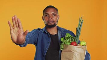 Vegan person showing a stop sign with his palm while he carries a bag full of groceries from local farmers market. Young adult restricting the access, expressing rejection in studio. Camera A. video
