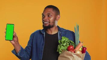 African american man showing a phone with isolated mockup screen, carrying a paper bag full of ethically sourced produce in studio. Model presenting greenscreen and eco friendly food. Camera A. video