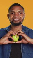 vertical africano americano relajado chico lo hace un corazón forma firmar con un manzana, recomendando en la zona crecido Produce en estudio. contento satisfecho persona disfruta sano comiendo y productos quimicos gratis nutrición. cámara una. video