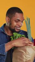 Vertikale vegan männlich Person bewundern frisch geerntet Früchte und Gemüse gekauft von lokal Bauern Markt, Orange Hintergrund. jung Erwachsene Gefühl zufrieden mit seine Tasche voll von organisch Waren. Kamera a. video