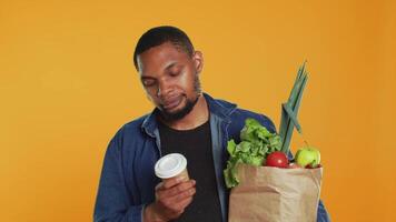 Young adult enjoying a cup of coffee during his grocery shopping spree, carrying a paper bag full of organic produce in studio. Man serving caffeine refreshment, zero waste. Camera A. video