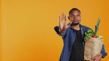 Vegan person showing a stop sign with his palm while he carries a bag full of groceries from local farmers market. Young adult restricting the access, expressing rejection in studio. Camera B. video