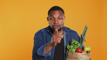 African american secretive guy doing mute hush gesture with finger over lips, keeping a secret about locally grown organic produce in a paper bag. Person being private about veganism. Camera A. video