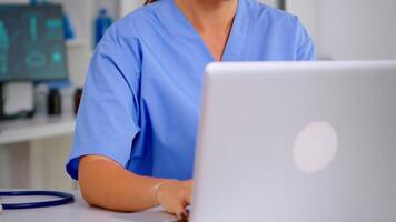 Close up of young medical assistant typing at laptop and raising head smiling at camera sitting in hospital office wearing blue uniform. Hospital healthcare worker, medicine clinical assistant health consultant therapist video