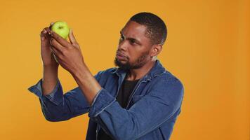 ung man inspekterande en nyligen UPPTAGITS grön äpple i studio, framställning Säker dess rena och mogen för förbrukande. vegan person undersöker organisk frukt från lokal- noll avfall eco Lagra. kamera a. video