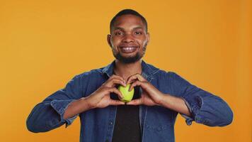 African american relaxed guy does a heart shape sign with an apple, recommending locally grown produce in studio. Happy pleased person enjoys healthy eating and chemicals free nutrition. Camera A. video