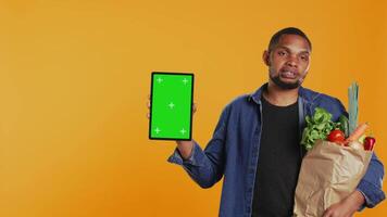 Vegan adult holding a tablet with greenscreen layout in studio, carrying paper bag full of ethically sourced chemicals free produce. Guy advocating for healthy eating with mockup screen. Camera B. video