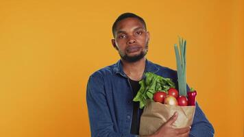 porträtt av afrikansk amerikan kille innehav en papper väska full av bio mat, uppköp organisk lokalt vuxen frukt och grönsaker. självsäker vegan person stödjande friska äter och noll avfall. kamera a. video