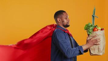 African american guy posing as a superhero with fresh ripe produce, hero with red cape promoting ethically sourced food and zero waste. Male model supports local farming. Camera B. video