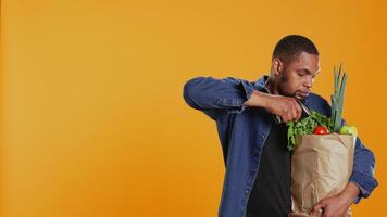 Young person scanning fresh organic produce in a paper bag, using scanner at local supermarket checkout. Male model carrying freshly harvested fruits and vegetables. Camera B. video